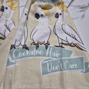 May include: Two beige kitchen towels with a blue crocheted trim and a button closure. Each towel features a repeating design of white cockatoos with yellow crests and the text "Cockatoo Hair Don't Care" in a banner.