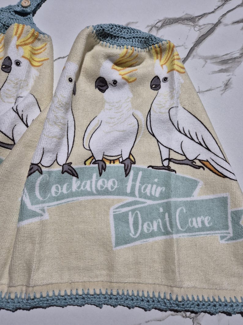 May include: Two beige kitchen towels with a blue crocheted trim and a button closure. Each towel features a repeating design of white cockatoos with yellow crests and the text "Cockatoo Hair Don't Care" in a banner.