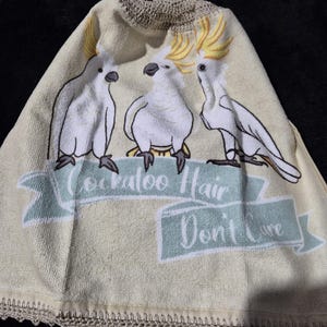 May include: A light beige towel with three white cockatoo bird illustrations and yellow crests. The towel has a light blue banner with the text "Cockatoo Hair Don't Care". The bottom edge has a decorative crochet trim.