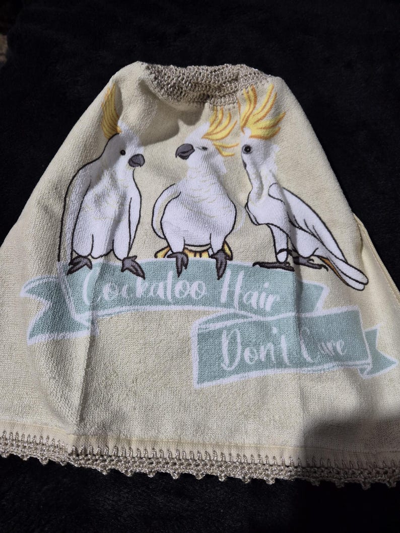 May include: A light beige towel with three white cockatoo bird illustrations and yellow crests. The towel has a light blue banner with the text "Cockatoo Hair Don't Care". The bottom edge has a decorative crochet trim.