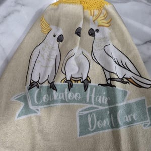 May include: A cream-colored kitchen towel featuring three white cockatoos with yellow crests. The towel has a yellow crocheted trim and a loop for hanging. Text on the towel reads "Cockatoo Hair Don't Care."