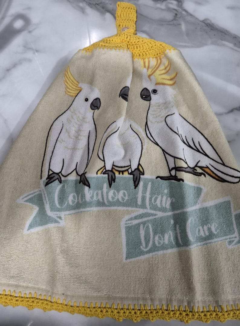 May include: A cream-colored kitchen towel featuring three white cockatoos with yellow crests. The towel has a yellow crocheted trim and a loop for hanging. Text on the towel reads "Cockatoo Hair Don't Care."