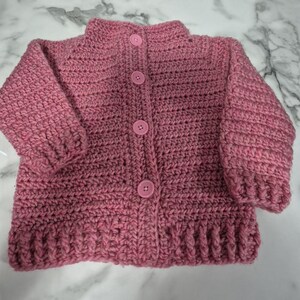 May include: A pink crocheted cardigan with a button closure. The cardigan has a ribbed texture and a high neckline.