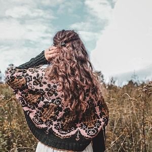 Può includere: Una donna con lunghi capelli castani ricci indossa un maglione con motivi neri, rosa e dorati. È in piedi in un campo di erba alta con un cielo nuvoloso sullo sfondo.