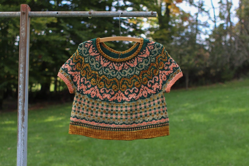 May include: A green, pink and gold knitted sweater with a short sleeve design. The sweater has a geometric pattern and is hanging on a wooden hanger.