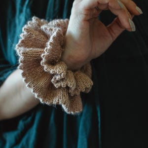 Könnte beinhalten: Ein gestricktes Armband in hellbrauner Farbe. Die Manschette hat ein gerüschtes, mehrschichtiges Design und wird am Handgelenk getragen. Der Hintergrund ist dunkelblaugrün.