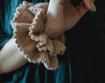 May include: A knitted cuff bracelet in a light brown color. The cuff has a ruffled, layered design, and is worn on the wrist. The background is a dark teal color.