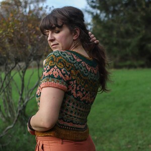 May include: A woman wearing a short-sleeved knitted jumper with a colourful geometric pattern in shades of green, pink, and gold. The jumper has a crew neckline and is cropped.
