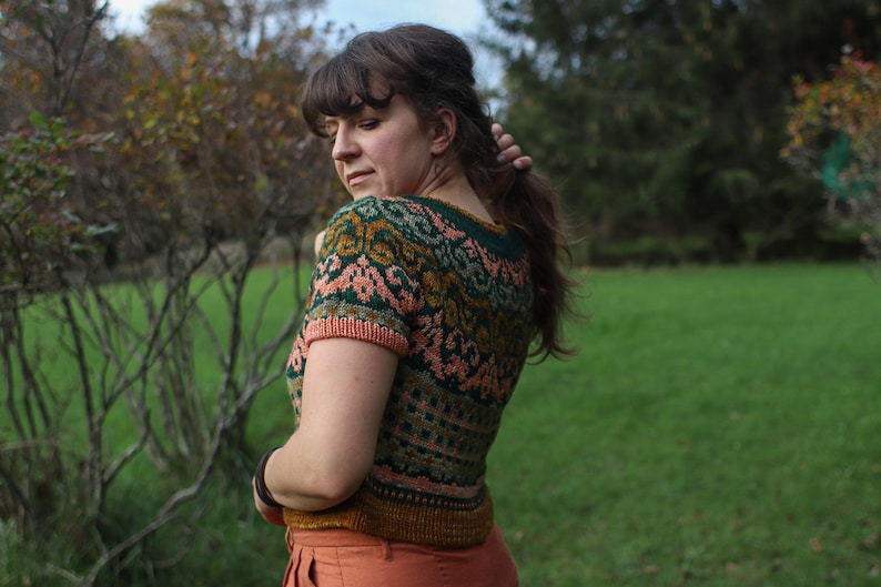 May include: A woman wearing a short-sleeved knitted jumper with a colourful geometric pattern in shades of green, pink, and gold. The jumper has a crew neckline and is cropped.