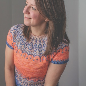 Op de afbeelding: Een vrouw draagt een gebreide trui met korte mouwen met een kleurrijk geometrisch patroon in tinten blauw, roze en oranje.