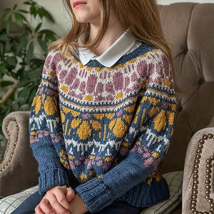 以下が含まれることがあります： 青、黄、ピンク、白のフェアアイル柄のニットセーター。セーターは丸首で長袖です。