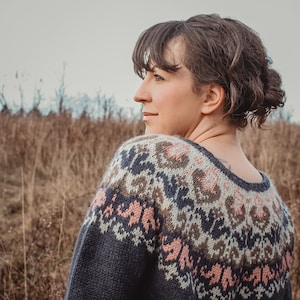 May include: A woman wearing a blue, brown, and pink patterned sweater with a fair isle design. She is standing in a field of tall, dry grass.