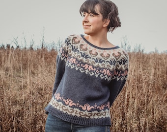 May include: A woman wearing a blue sweater with a pink, brown, and white patterned design. She is standing in a field of tall, dry grass.