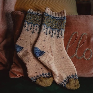 Pode incluir: Um par de meias feitas à mão em cor pêssego claro com detalhes estampados em azul e verde. As meias têm calcanhares azuis e dedos verde-oliva. A parte superior das meias tem uma borda ondulada.