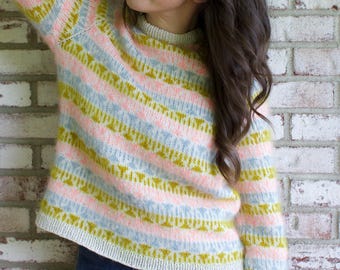 May include: A striped sweater in pastel colors of pink, blue, and yellow. The sweater has long sleeves and a crew neck. The person in the image is wearing blue jeans and leaning against a brick wall.
