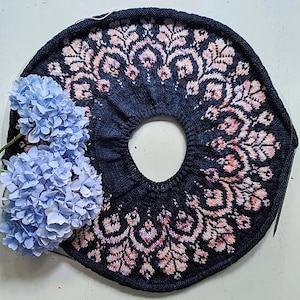 以下が含まれることがあります： 白い背景の上に置かれた、濃い青色の編まれた円形。縁にはピンクと白の花柄が施されています。