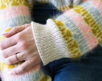 May include: A cozy, striped sweater in pastel colors. The sweater features horizontal stripes of pink, yellow, and blue. The cuffs are a solid cream color. The sweater is made of a soft, fuzzy material.
