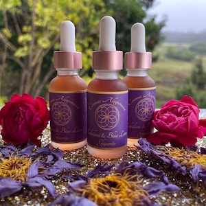 May include: Three small glass bottles with dropper tops filled with a light brown liquid. The bottles have purple labels with gold lettering that reads "Cosmic Alignment" and "Celandine & Blue Lotus Glycerite". The bottles are sitting on a gold sparkly surface with dried flowers.