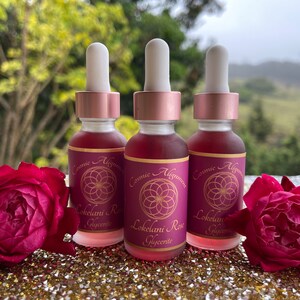 May include: Three glass bottles with dropper tops filled with a pink liquid. The bottles have labels with the text "Cosmic Alignment" and "Lokelani Rose Glycerite". The bottles are sitting on a sparkly gold surface with two red roses.