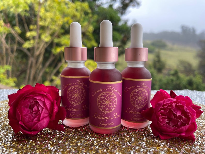 May include: Three glass bottles with dropper tops filled with a pink liquid. The bottles have labels with the text "Cosmic Alignment" and "Lokelani Rose Glycerite". The bottles are sitting on a sparkly gold surface with two red roses.
