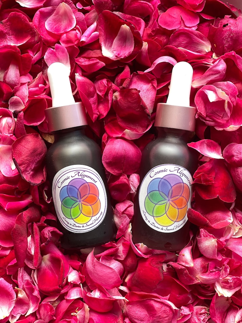 May include: Two dark brown glass bottles with dropper tops and white labels. The labels have a colorful mandala design and the text "Cosmic Alignment Living in Divine & Sacred Harmony". The bottles are surrounded by pink rose petals.
