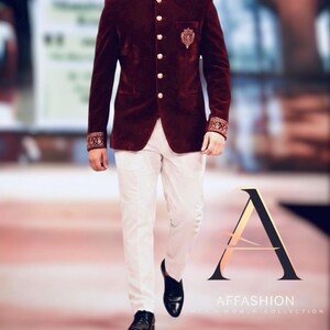 K&ouml;nnte beinhalten: Burgunderfarbene Samtjacke mit goldenen Kn&ouml;pfen und bestickten Manschetten, kombiniert mit wei&szlig;er Hose und schwarzen Schuhen. Die Jacke hat ein verziertes Emblem auf der Brust. Der Text "AFFASHION MEN'S WOMEN COLLECTION" ist enthalten.