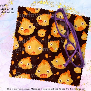 May include: A 6x7 inch microfiber cleaning cloth with a black background and a repeating pattern of cartoon fire spirits with orange flames. The cloth is folded and a purple pair of glasses is resting on top of it.