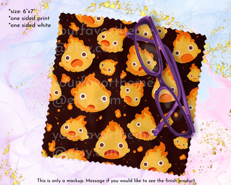 May include: A 6x7 inch microfiber cleaning cloth with a black background and a repeating pattern of cartoon fire spirits with orange flames. The cloth is folded and a purple pair of glasses is resting on top of it.