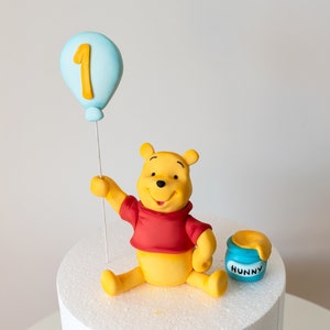 Può includere: Un topper per torta di Winnie the Pooh con un palloncino blu su cui è scritto "1". Winnie the Pooh tiene il palloncino e ha un barattolo di miele su cui è scritto "Hunny".