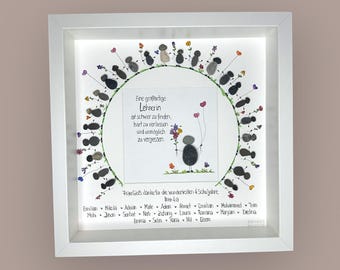May include: A framed artwork with a white border, featuring a pebble art design. The artwork includes a central circular design with text in German and pebble figures. The frame is square and sits on a neutral background.
