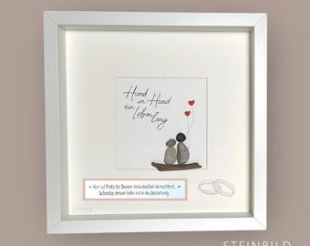 May include: A framed artwork featuring a pebble art scene of two figures holding hands, with three red heart balloons. The text "Hand in Hand ein Lebenlang" is written above. Two silver rings are depicted below the artwork.