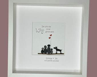 May include: A square white framed wall art piece featuring pebble art figures of four people and a dog standing on a twig. Above them, two red heart shapes are drawn. The text reads: Der schönste Weg ist der gemeinsame, Dominique ♥ Kim mit Charlotte und Jonah.