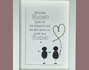May include: A white framed wall art piece featuring two figures made of black stones holding hands. Above them, the German text reads: Wir bleiben FREUNDINNEN bis wir alt und vergesslich sind und dann werden wir wieder neue FREUNDINNEN. A heart shape made of black and white twine is attached.