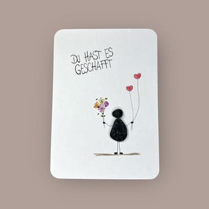 Puede incluir: Una tarjeta blanca con esquinas redondeadas presenta una figura de palo sosteniendo flores y dos globos en forma de corazón. El texto "DU HAST ES GESCHAFFT" está escrito en negro en la parte superior. La figura está hecha de guijarros negros.