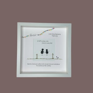 May include: A framed artwork with a white border. The artwork features a drawing of two figures on a line, with text in German. The text includes the name "Alexander" and the date "11. April 2026". Colorful bunting decorates the top.