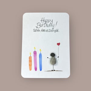 以下が含まれることがあります： 「Happy Birthday!」と「Schön, dass es Dich gibt.」の文字が入った白いバースデーカード。石でできた棒人間が赤いハートの風船を持っています。フィギュアの左側には3本のカラフルなろうそくがあります。