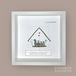 Könnte beinhalten: Ein weißer Rahmen mit einem weißen Passepartout und einem weißen Hintergrund. Der Rahmen enthält ein Steinbild von einem Haus mit einer fünfköpfigen Familie. Die Namen Tim, Minam, Nede und Mia sind auf dem Haus geschrieben. Der Text "Familie Dieckerhoff" befindet sich oben im Rahmen. Der Text "Hier ist Platz für Deinen individuellen Wunschtext. Schreibe diesen bitte mit in die Bestellung." befindet sich unten im Rahmen.