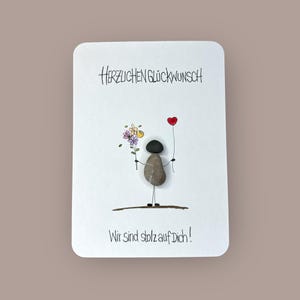 May include: A white card with the text "HERZLICHEN GLÜCKWUNSCH" and "Wir sind stolz auf Dich!" features a pebble art figure holding flowers and a red heart balloon. The figure stands on a brown line, with a light brown background.