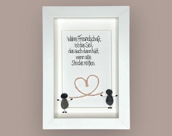 May include: A framed artwork with a white border. The artwork features the text "Wahre Freundschaft ist das Seil, das auch dann hält, wenn alle Stricke reißen." in black script. Below the text is a heart shape made of a light brown material, and two small figures.