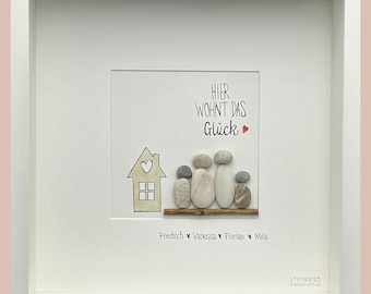 May include: A framed pebble art picture with a house and four figures representing a family. The text reads "Hier wohnt das Glück" which translates to "Here lives happiness". The names Friedrich, Vanessa, Florian, and Mila are written below the figures.