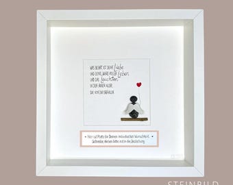 May include: White framed picture with a white background and a black and white pebble art angel holding a red heart. The text reads "WAS BLEIBT, IST DEINE Liebe UND DENE JAHRE VOLLER Leben UND DAS leuchten IN DEN AUGEN ALLER DIE VON DER ERZÄHLEN" and "Hier ist Platz für Deinen individuellen Wunschtext Schreibe diesen bitte nit in die Bestellung". The bottom right corner of the frame has the text "STEINBILD MANUFAKTUR".