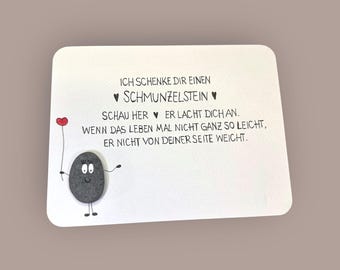 May include: A white card with German text and a cartoon illustration of a smiling stone holding a red heart balloon. The text reads, "I give you a Schmumzelstein. Look here, he laughs at you. When life is not so easy, he does not leave your side."