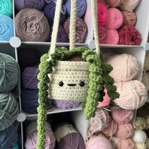 May include: A crocheted hanging planter with a green plant and a smiling face. The planter is hanging from a white cord and is surrounded by balls of yarn in various colors.