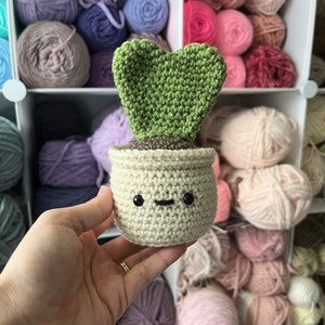 May include: A crocheted heart-shaped plant in a white pot with a cute face. The plant is green and the pot has black eyes and a smile. The plant is surrounded by balls of yarn in various colors.