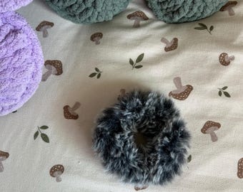 May include: A circular hair scrunchie made of faux fur with a black and grey mottled pattern. The scrunchie is positioned on a light-colored fabric featuring a repeating print of small brown mushrooms and green leaves.