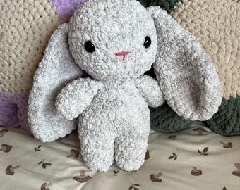 May include: A plush stuffed animal shaped like a rabbit, crafted from soft, light gray textured yarn. The rabbit features large, floppy ears, round black plastic eyes, and a small, hand-stitched pink nose and mouth detail.