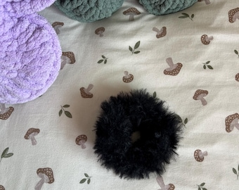 May include: A black faux fur hair scrunchie with a soft, fuzzy texture. The circular hair accessory is positioned on a light-colored fabric surface featuring a repeating pattern of small brown mushrooms and green leaves.