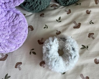 May include: A fluffy white and gray faux fur hair scrunchie with a soft, textured appearance. The scrunchie is circular with a gathered elastic center, featuring a mix of light gray and white fibers throughout its plush, shaggy surface.