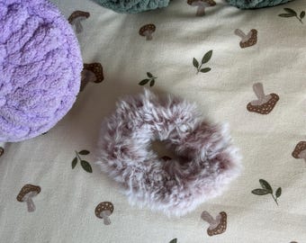 May include: A fluffy, mauve-colored faux fur hair scrunchie with a soft, textured appearance. The scrunchie is circular with a gathered elastic center, featuring long, synthetic fibers that create a dense, plush look.