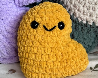 May include: A mustard yellow plush toy shaped like a heart with a soft, chunky knit texture. It features two round, black plastic safety eyes and a simple, hand-stitched black V-shaped mouth, giving it a friendly, smiling expression.